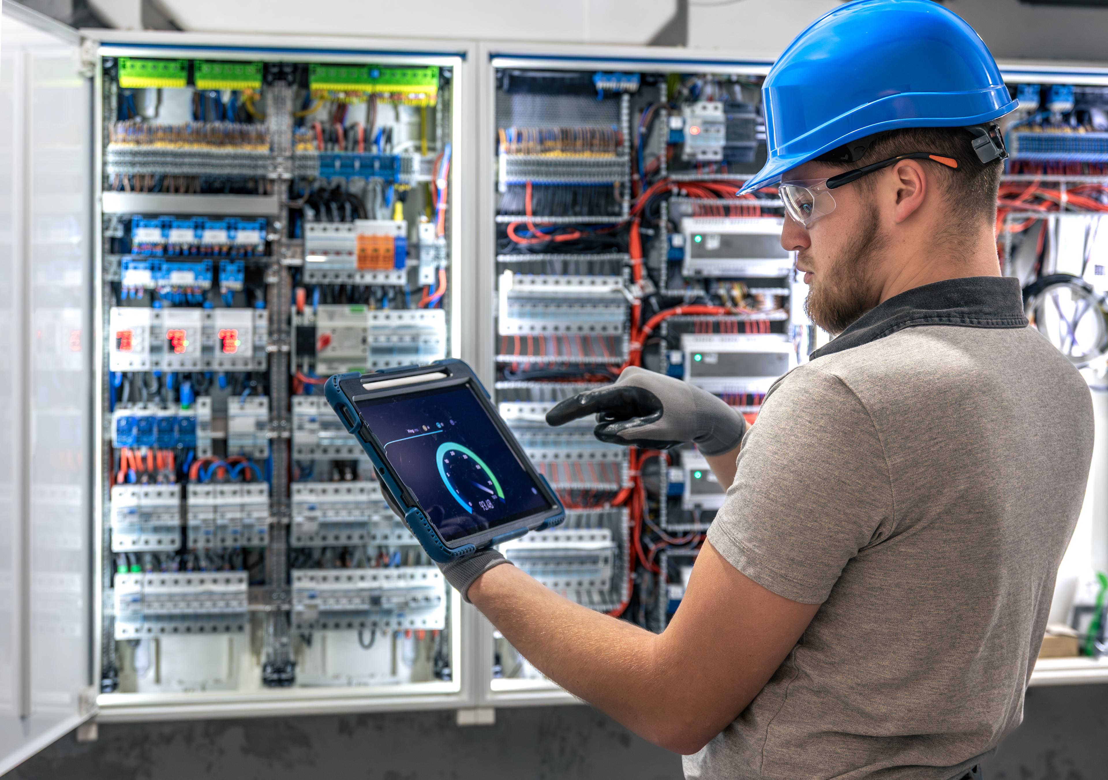 Electrician reviewing a digital system on a tablet at an industrial switchboard and control panel